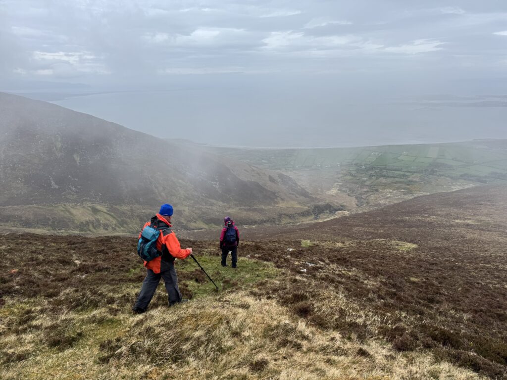 Two hikers descending through heather on the Caherconree and Baurtregaum Loop, with Derrymore Glen, green fields and the Kerry coastline coming back into view below the cloud.