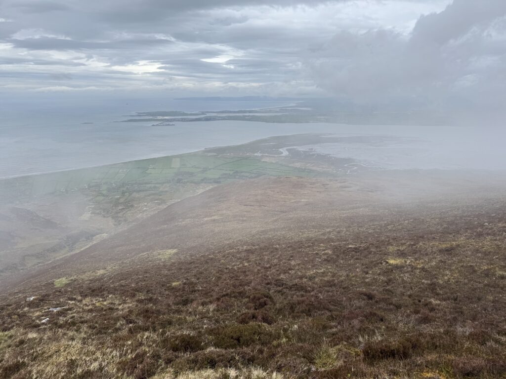 Wide view from the mountains over heather-covered slopes, patchwork fields and the coastline of Tralee Bay under dramatic cloud.