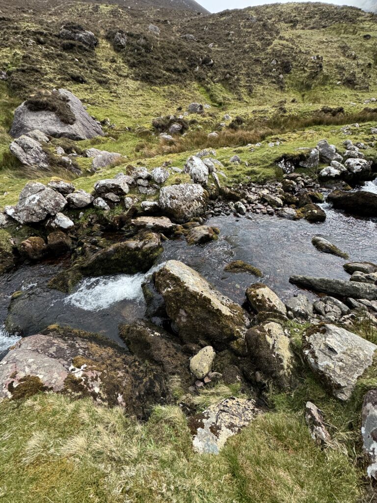 Rocky river crossing in Derrymore Glen, with fast-flowing water weaving between large boulders below grassy mountain slopes
