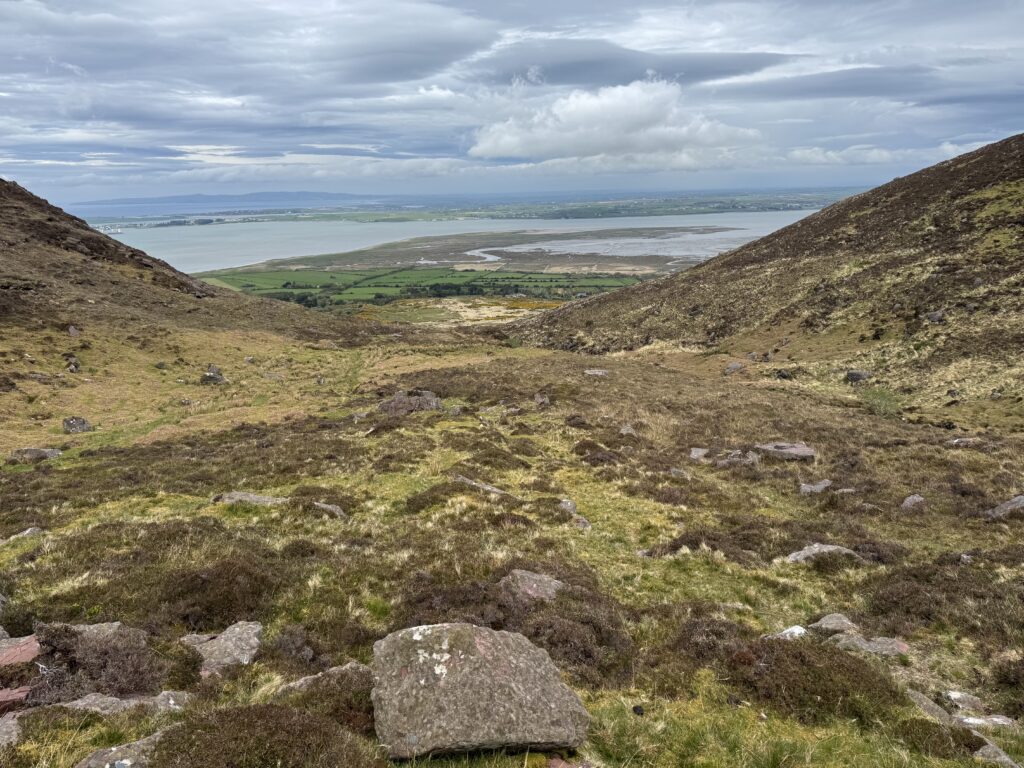 Wide mountain view through Derrymore Glen towards green farmland and the tidal flats of Tralee Bay, framed by steep slopes under broken cloud.
