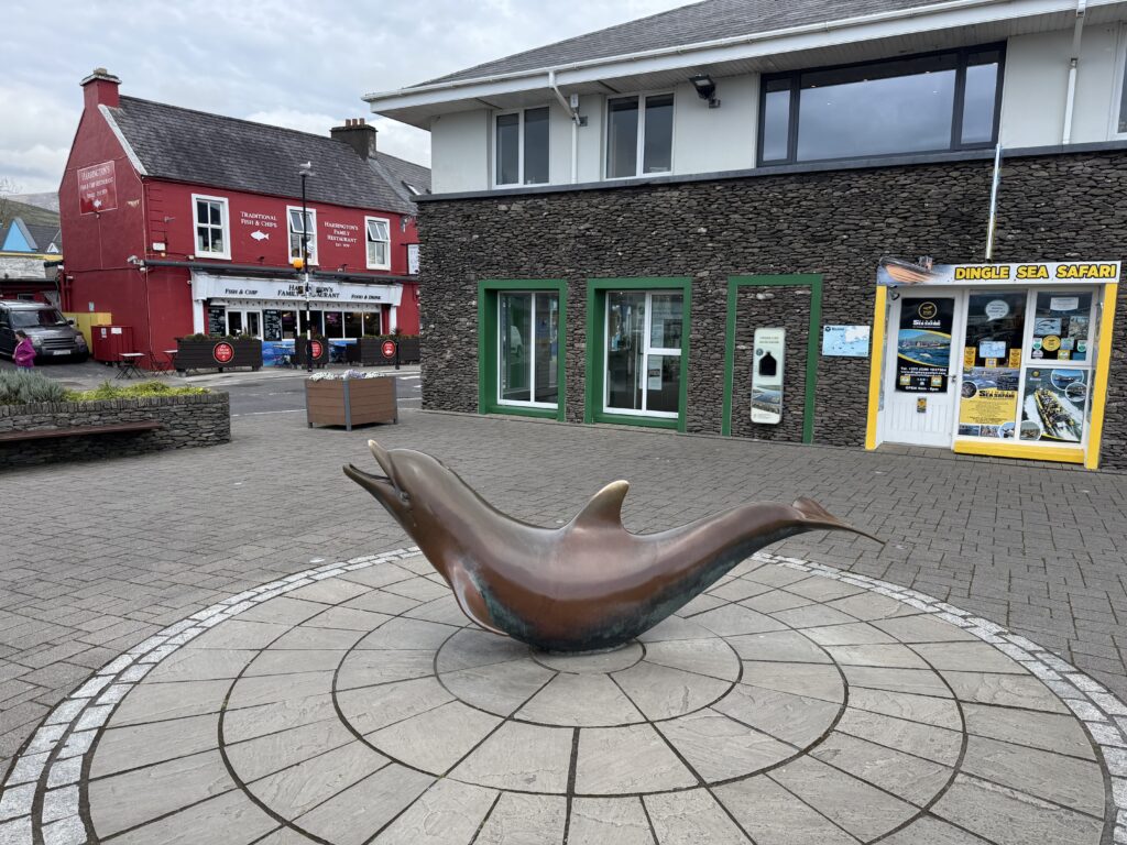 Bronze statue of Fungie the dolphin in Dingle town, County Kerry, displayed on a circular stone-paved area near the harbour, with a red fish and chip restaurant and local tour operators in the background.