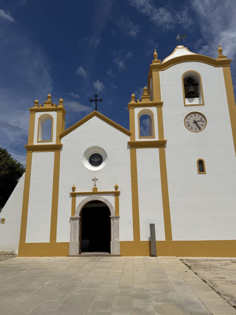 Beautiful whitewashed church in Praia da Luz, Portugal, featuring mustard-yellow architectural details, an arched entrance, circular window, bell tower and clock beneath a deep blue sky with scattered clouds.
