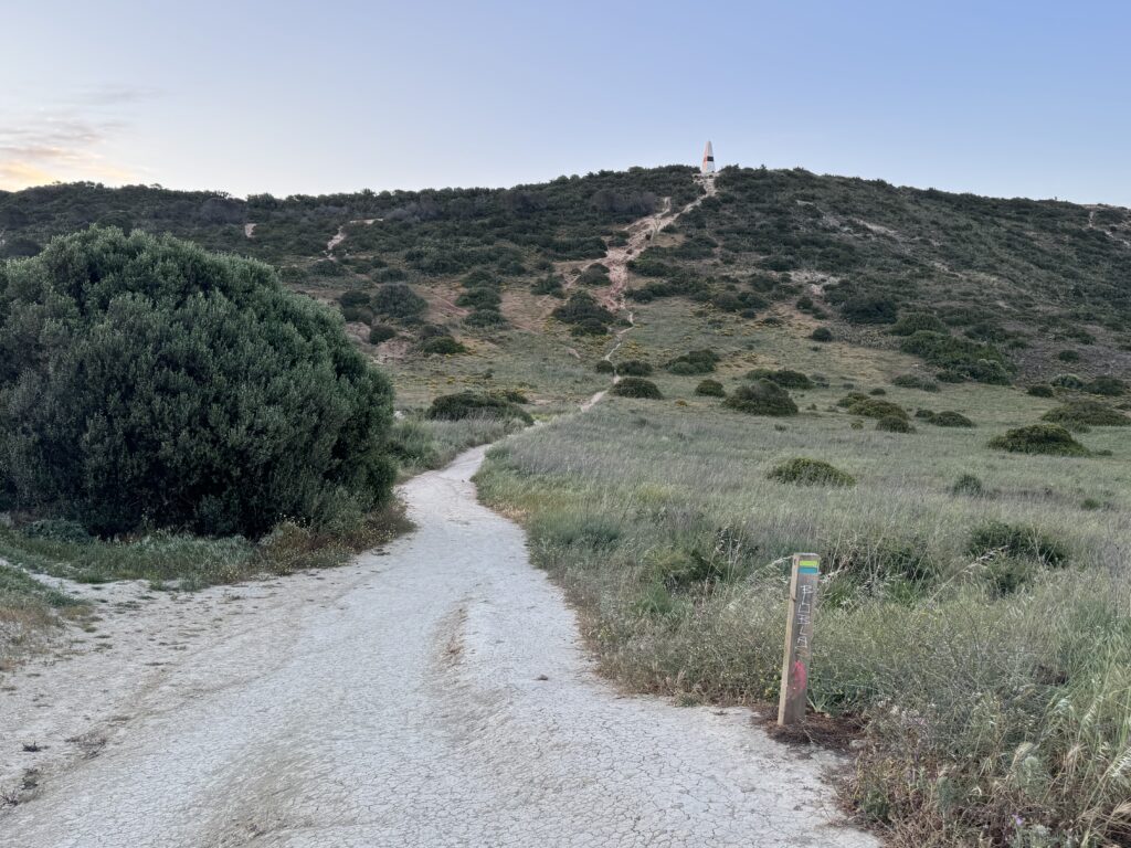 
A dusty walking trail winding through grassy slopes and low green shrubs towards a white obelisk with a black stripe on a hilltop near Praia da Luz, Portugal, with a waymarked trail post in the foreground.