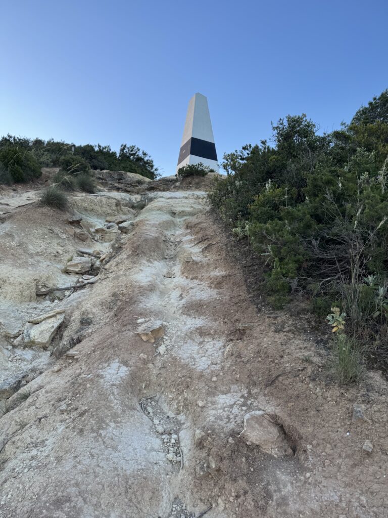 Close-up view of a steep, uneven hiking trail with exposed pale rock, loose stones and deep grooves, climbing towards a white obelisk with a black band, surrounded by dense green shrubs beneath a clear blue sky.