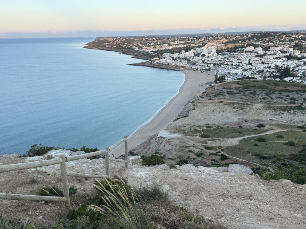 Elevated coastal view overlooking the curved sandy beach of Praia da Luz, with whitewashed buildings lining the shoreline, winding walking trails across the headland and the calm Atlantic Ocean beneath a pale evening sky.