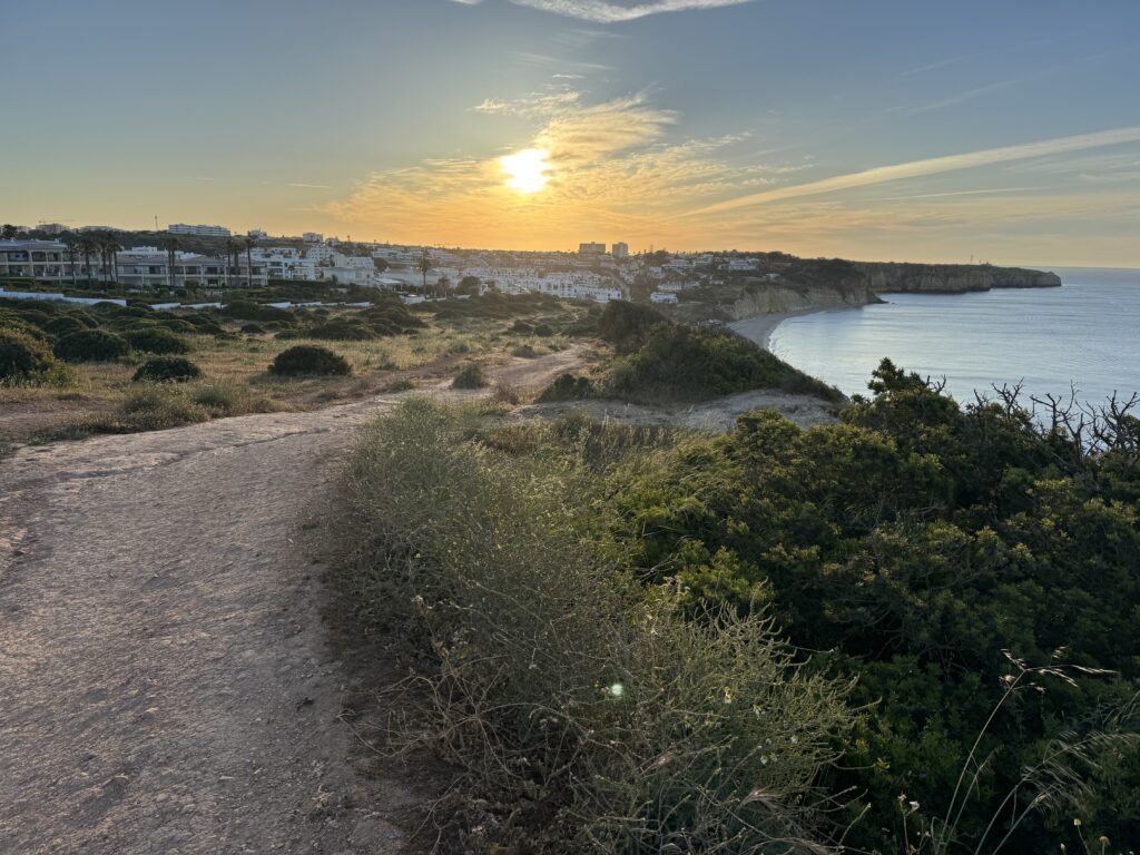 Scenic coastal walking trail above Praia da Luz in Portugal, with sandy paths winding through low green shrubs, overlooking the curved beach, whitewashed town and calm Atlantic Ocean as the sun sets over the coastline.