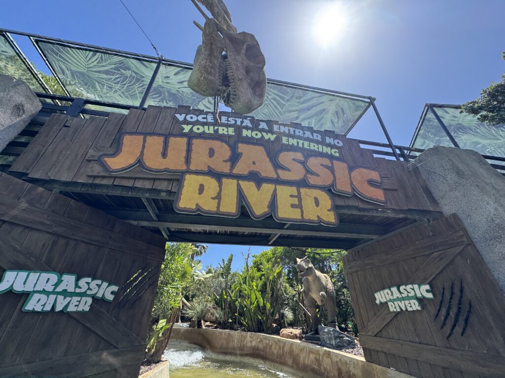 Wooden entrance gateway to the Jurassic River attraction at Zoomarine in Portugal, featuring a large dinosaur skull overhead, colourful lettering, tropical plants, a dinosaur model and a water channel passing beneath the entrance.