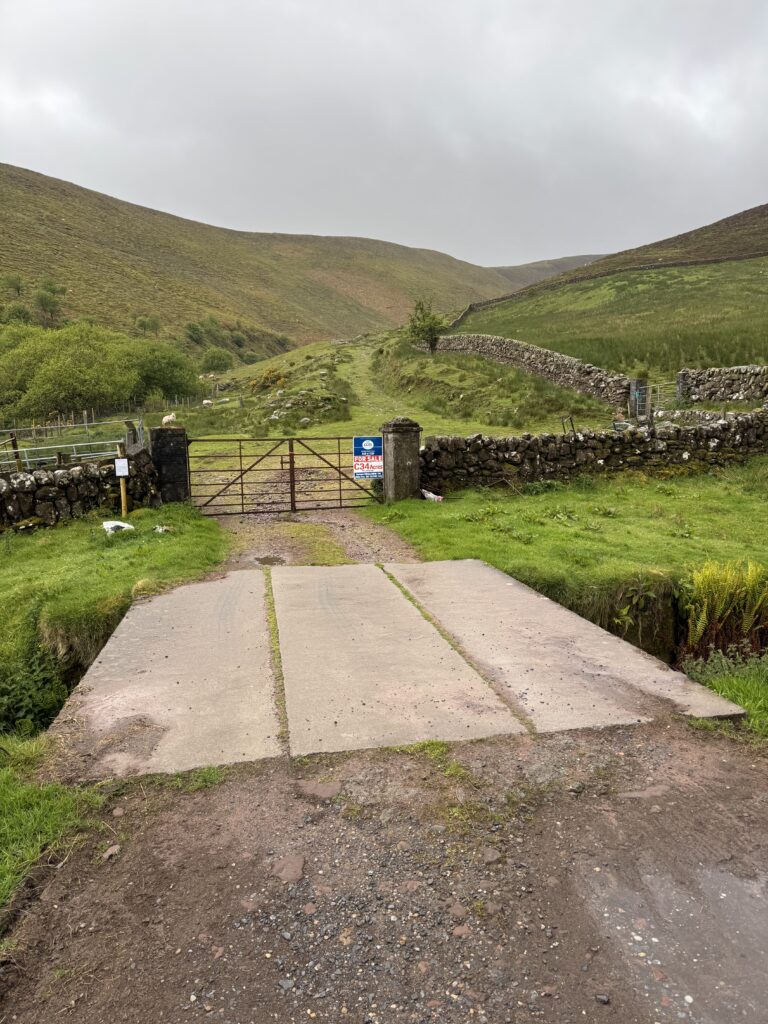 Small concrete bridge and farm gate at the entrance to a grassy mountain track, with dry-stone walls climbing through the valley under an overcast sky.