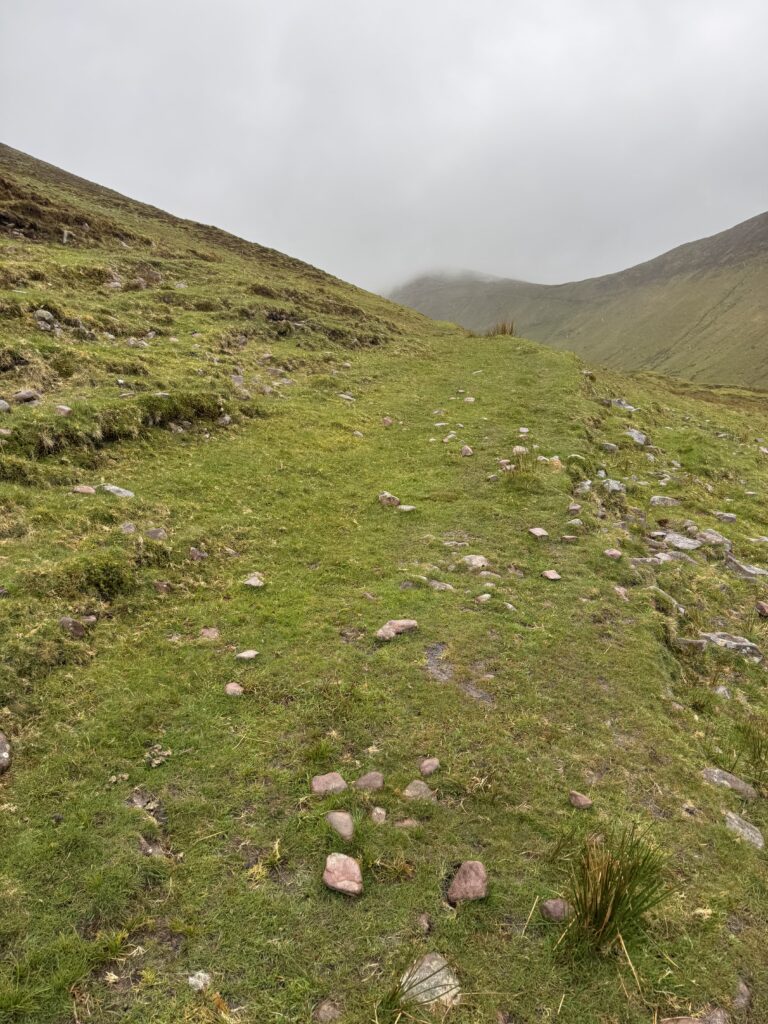 Faint grassy path climbing across an open mountainside towards a cloud-covered ridge, with scattered rocks and broad slopes in the Galtees.