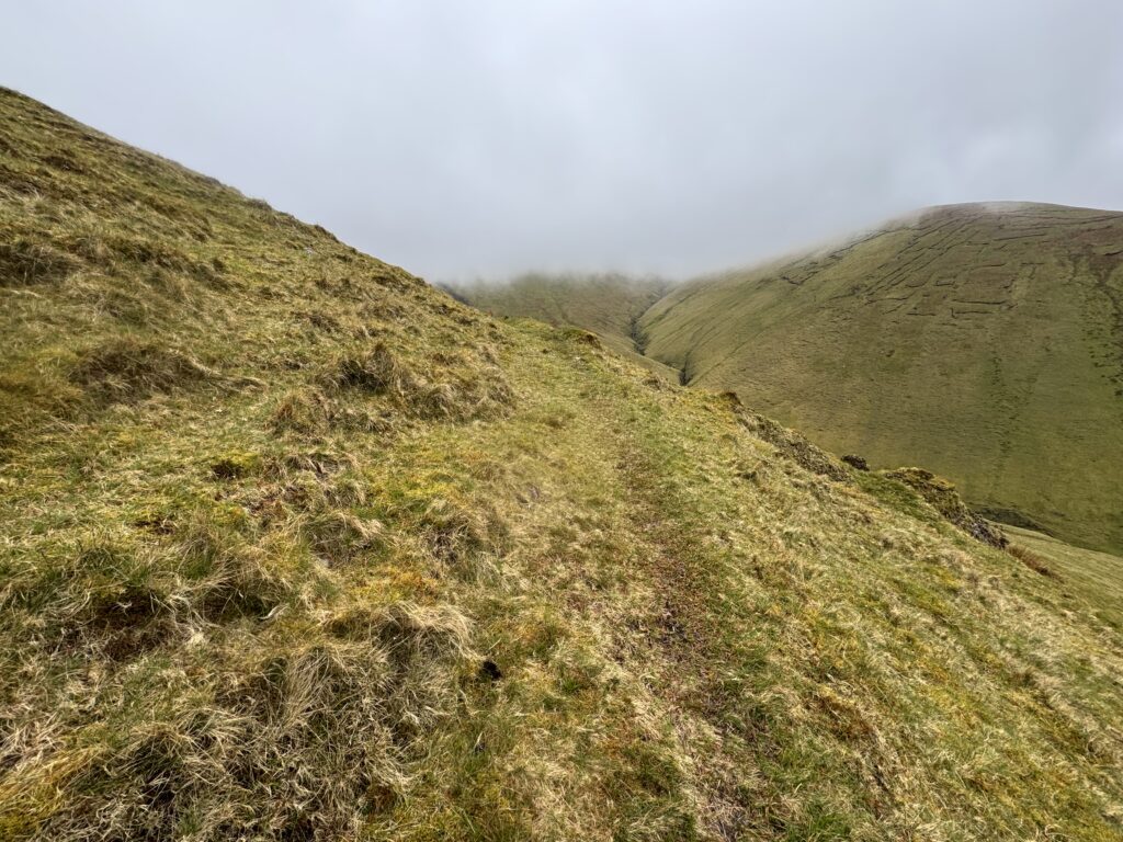 Narrow grassy trail on the Lyracappul Loop traversing a steep mountainside, with low cloud drifting across the rounded Galtee slopes opposite.