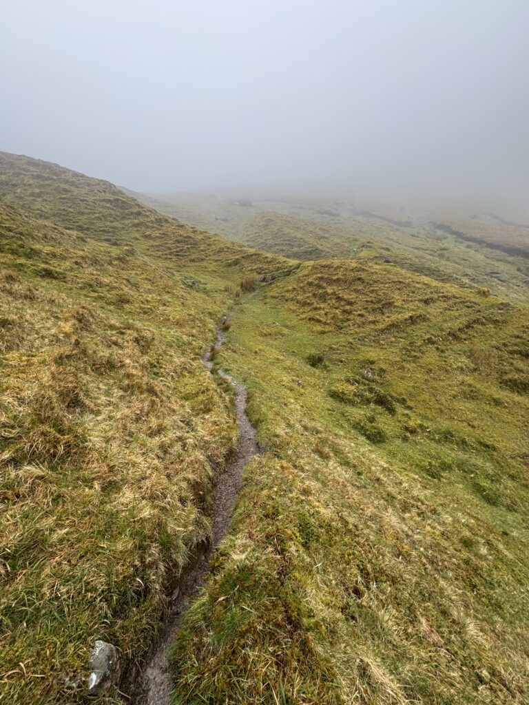 Faint mountain path winding through uneven grassy ground and disappearing into dense mist on the higher slopes of the Galtees.