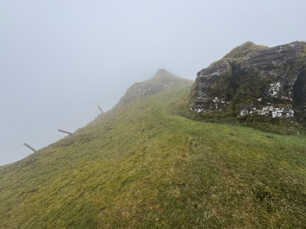 Narrow ridge section on the Lyracappul Loop, with grassy ground, rocky outcrops and steep drops disappearing into thick cloud in the Galtee Mountains.