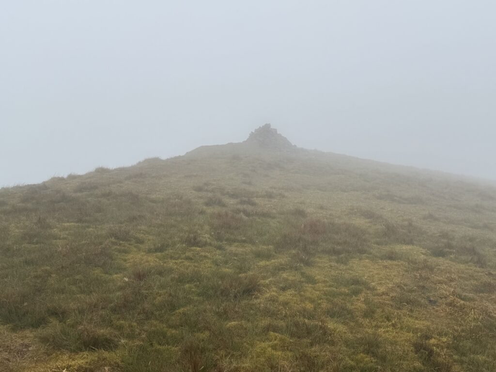 Stone summit cairn emerging through thick mist on the Lyracappul Loop, surrounded by rough grassy terrain on the upper slopes of the Galtee Mountains.