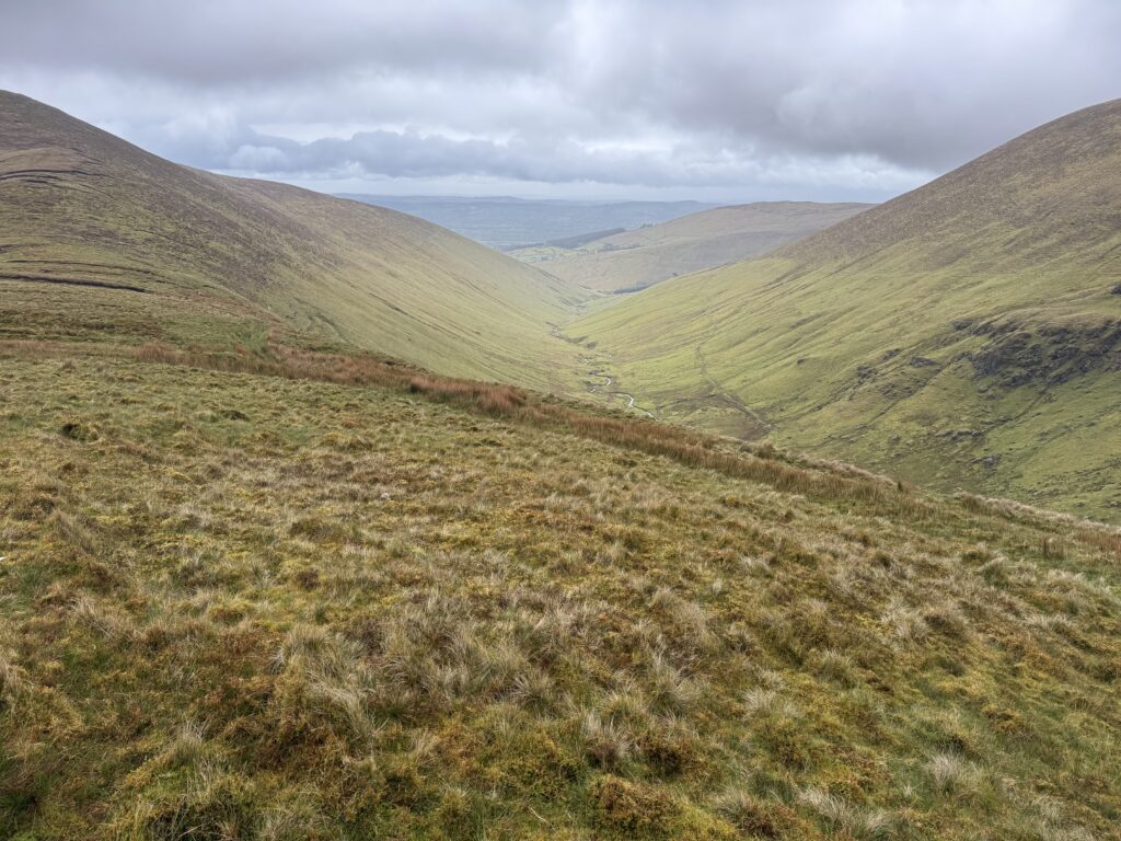 Broad green mountain valley with a winding stream on the valley floor, steep rounded slopes on either side and dark cloud hanging overhead.