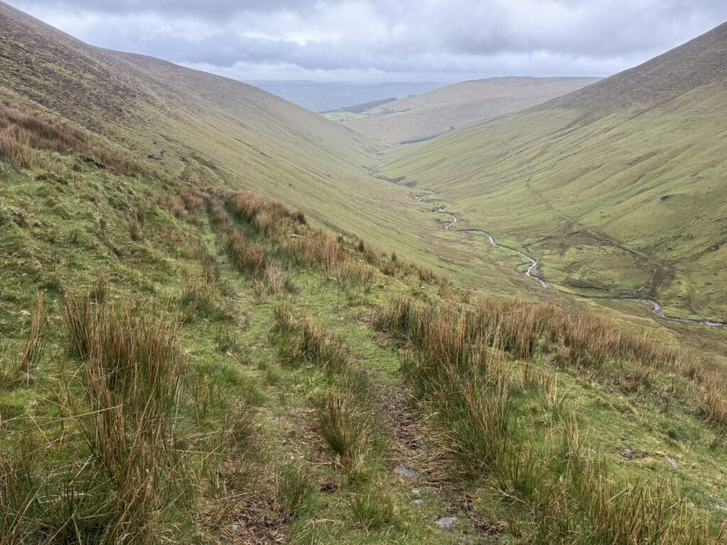 Grassy descent path above a wide mountain valley, with a winding river on the valley floor and rounded Galtee slopes beneath heavy cloud.