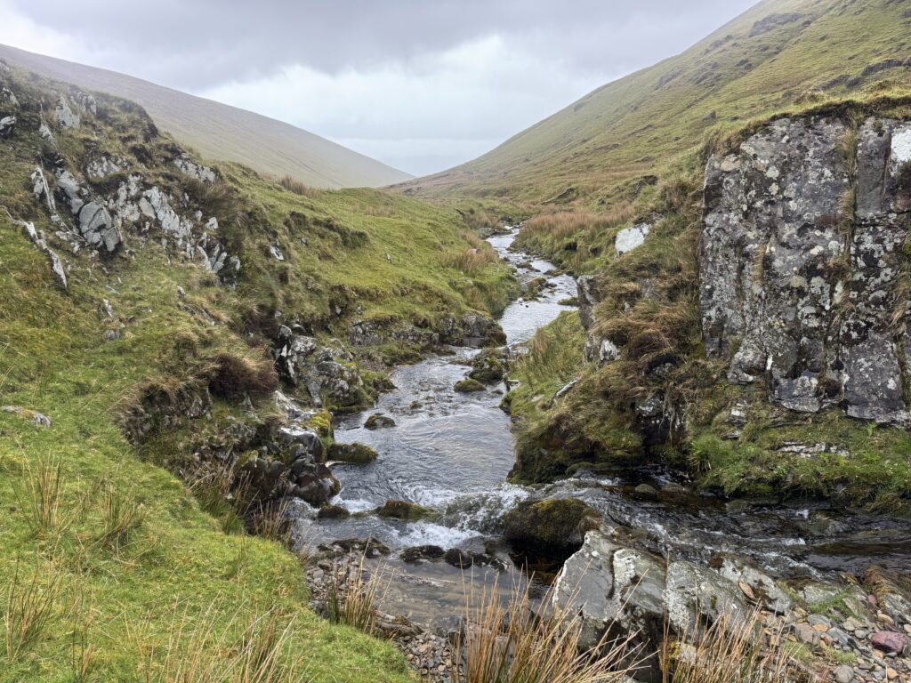 Rocky mountain stream on the Lyracappul Loop, flowing through a narrow grassy valley between steep slopes and exposed rock in the Galtee Mountains.