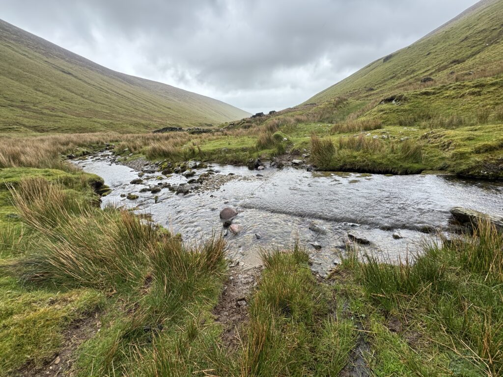 Fording point of a mountain river flowing across a raised concrete crossing, surrounded by grassy slopes and rushes beneath a cloudy sky.