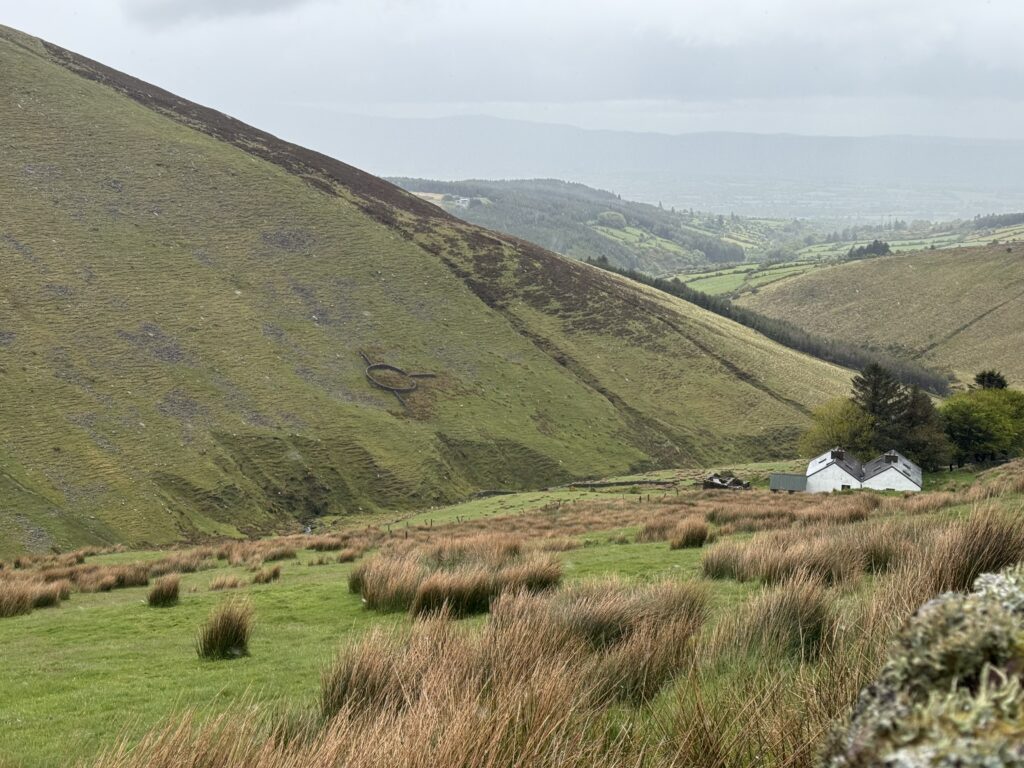 Remote white farmhouse beneath steep grassy mountain slopes, with a circular stone ringfort visible on the opposite hillside and farmland stretching into the distance.